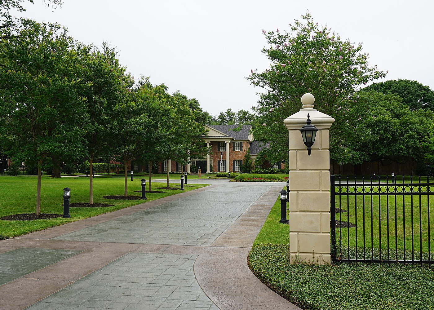 A landscaped entrance drive with stone gate posts and iron railings leading into a wooded residential property &mdash; representative imagery, not a specific Texas Pride job